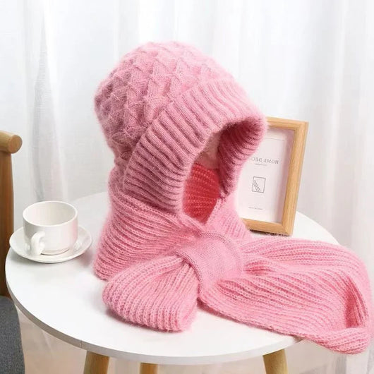Rosa Wintermütze mit Ohrenschutz und integriertem Schal auf einem weißen Tisch neben einer Kaffeetasse und einem Bilderrahmen.