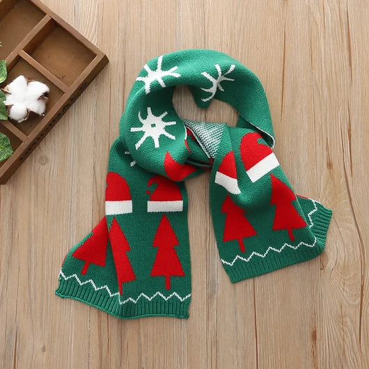 Grüner Kinderschal mit Weihnachtsmotiven wie Tannenbäumen, Weihnachtsmannmützen und Schneeflocken auf einem Holztisch.