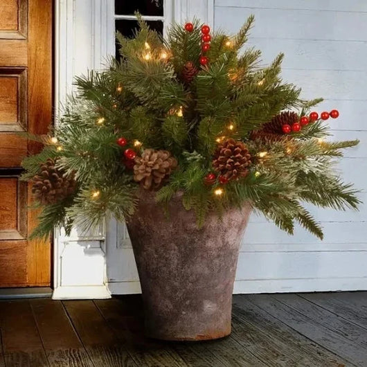 Ein dekorativer künstlicher Weihnachtsbaum in einem Tontopf mit Tannenzapfen, roten Beeren und einer Lichterkette.