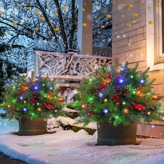 Zwei beleuchtete künstliche Weihnachtsbäume in Töpfen, dekoriert mit Tannenzapfen und Beeren, stehen im verschneiten Außenbereich.