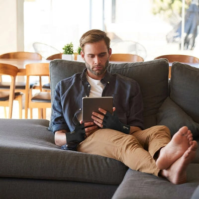 Ein Mann sitzt auf einem Sofa und trägt Kupferkompressionshandschuhe mit offenen Fingern, während er ein Tablet benutzt.