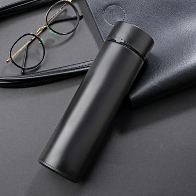 Schwarze Edelstahl-Thermosflasche mit LED-Temperaturanzeige neben einer Brille auf einer dunklen Oberfläche.