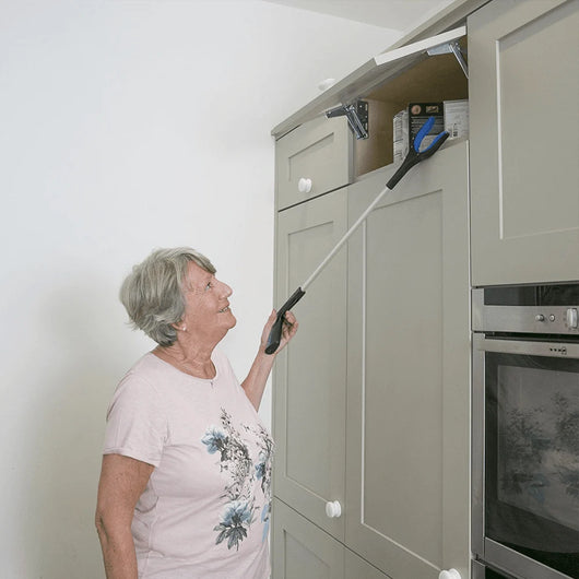 Eine ältere Frau benutzt einen faltbaren Greifer, um einen Gegenstand aus einem hochgelegenen Küchenschrank zu nehmen.
