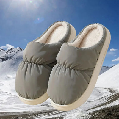 Ein Paar graue, gefütterte Hausschuhe vor einer verschneiten Berglandschaft.