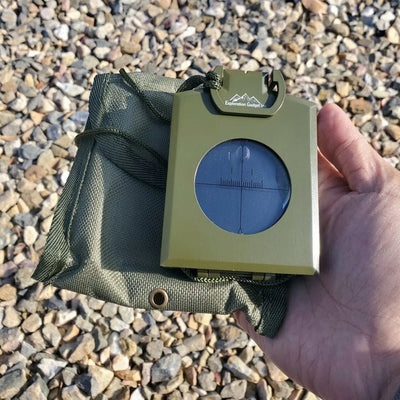 Ein olivgrüner Militärkompass mit Schutzhülle wird in einer Hand über einem steinigen Untergrund gehalten.